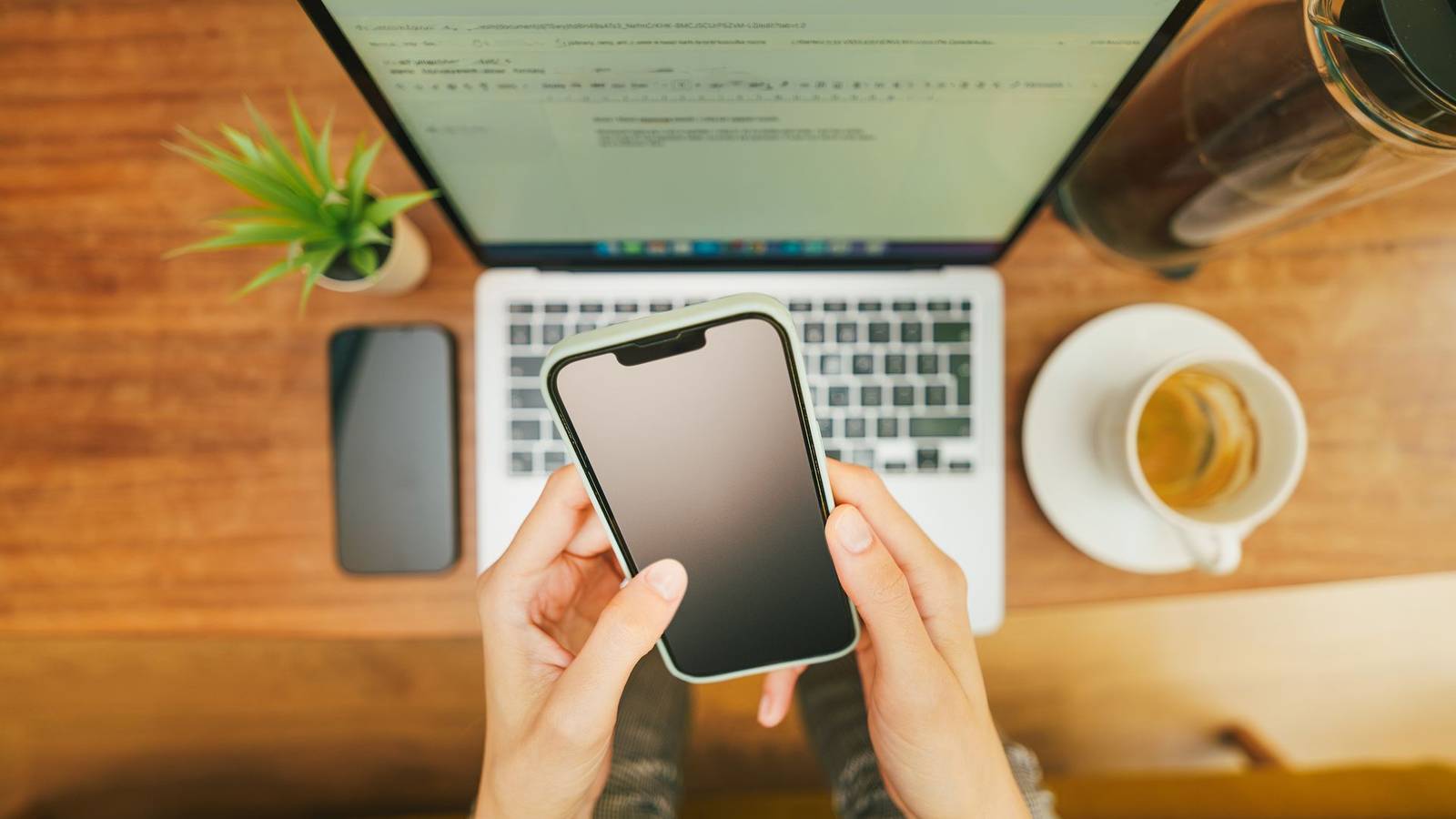 Hands holding a smartphone above a laptop on a wooden desk, with a cup of tea beside it