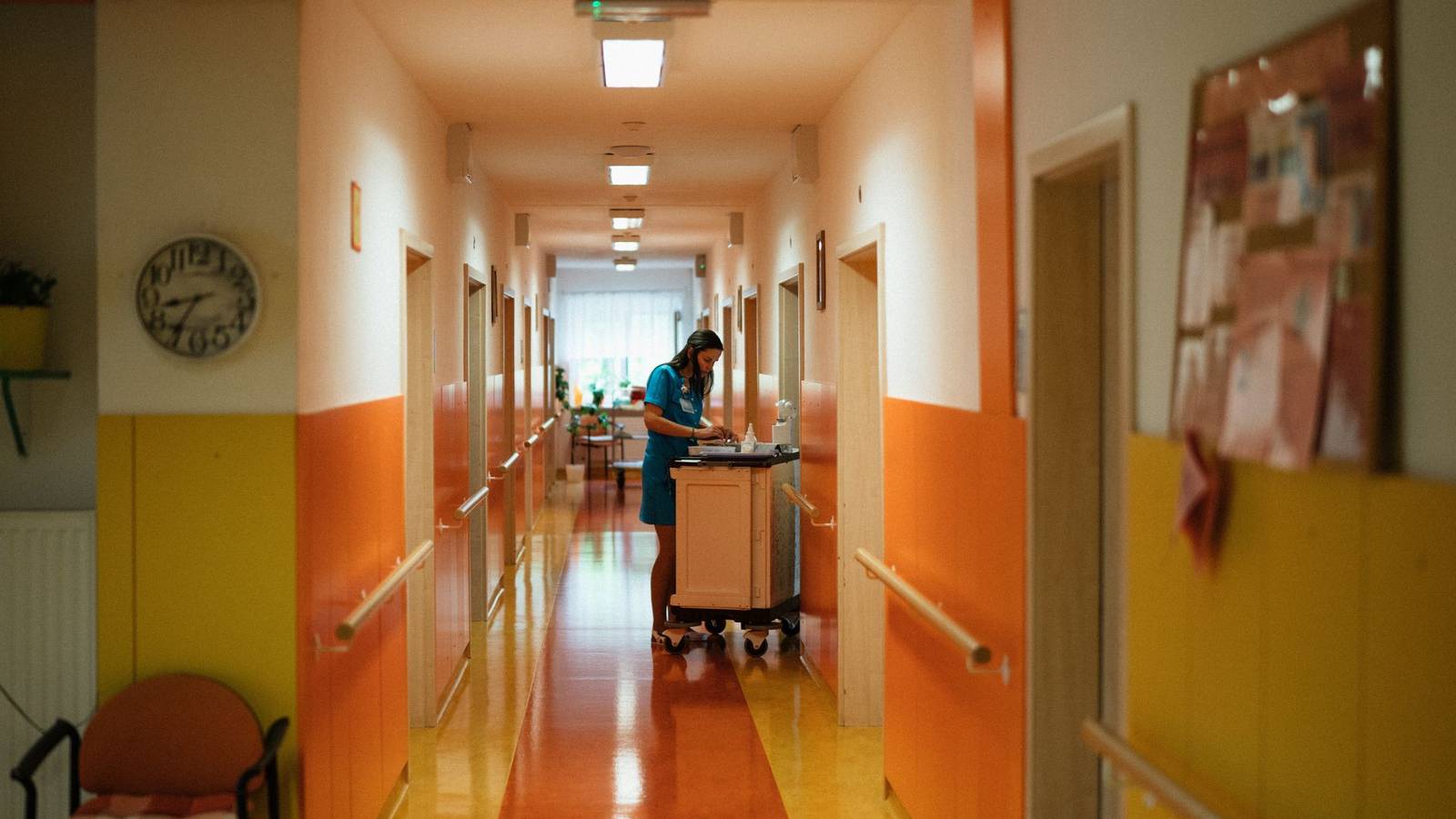 A nurse in teal scrubs works at a medicine trolley in a long, brightly painted hospital corridor