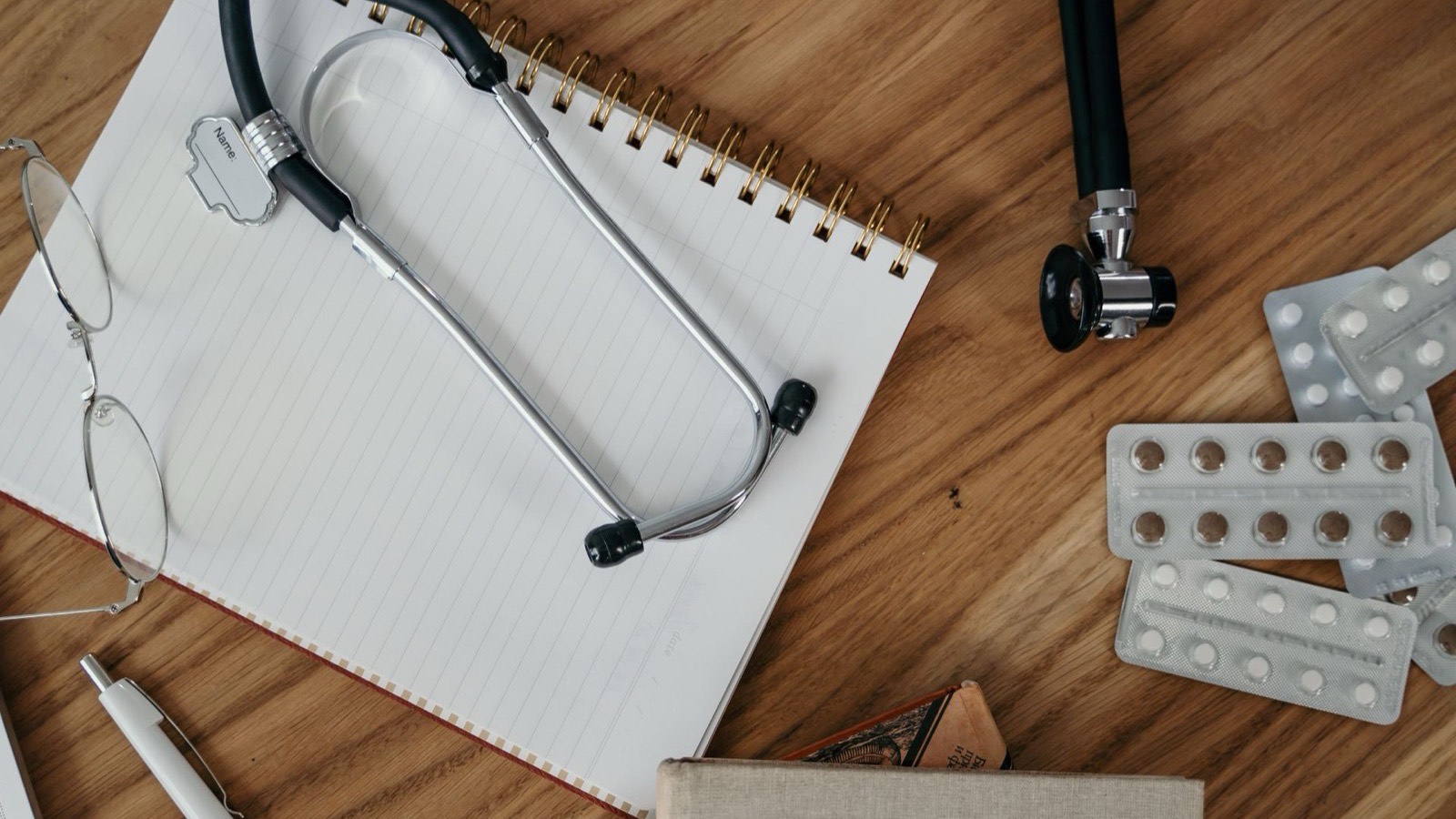 A stethoscope on an open spiral notebook, with glasses, a pen, books and blister packs of tablets on a wooden desk