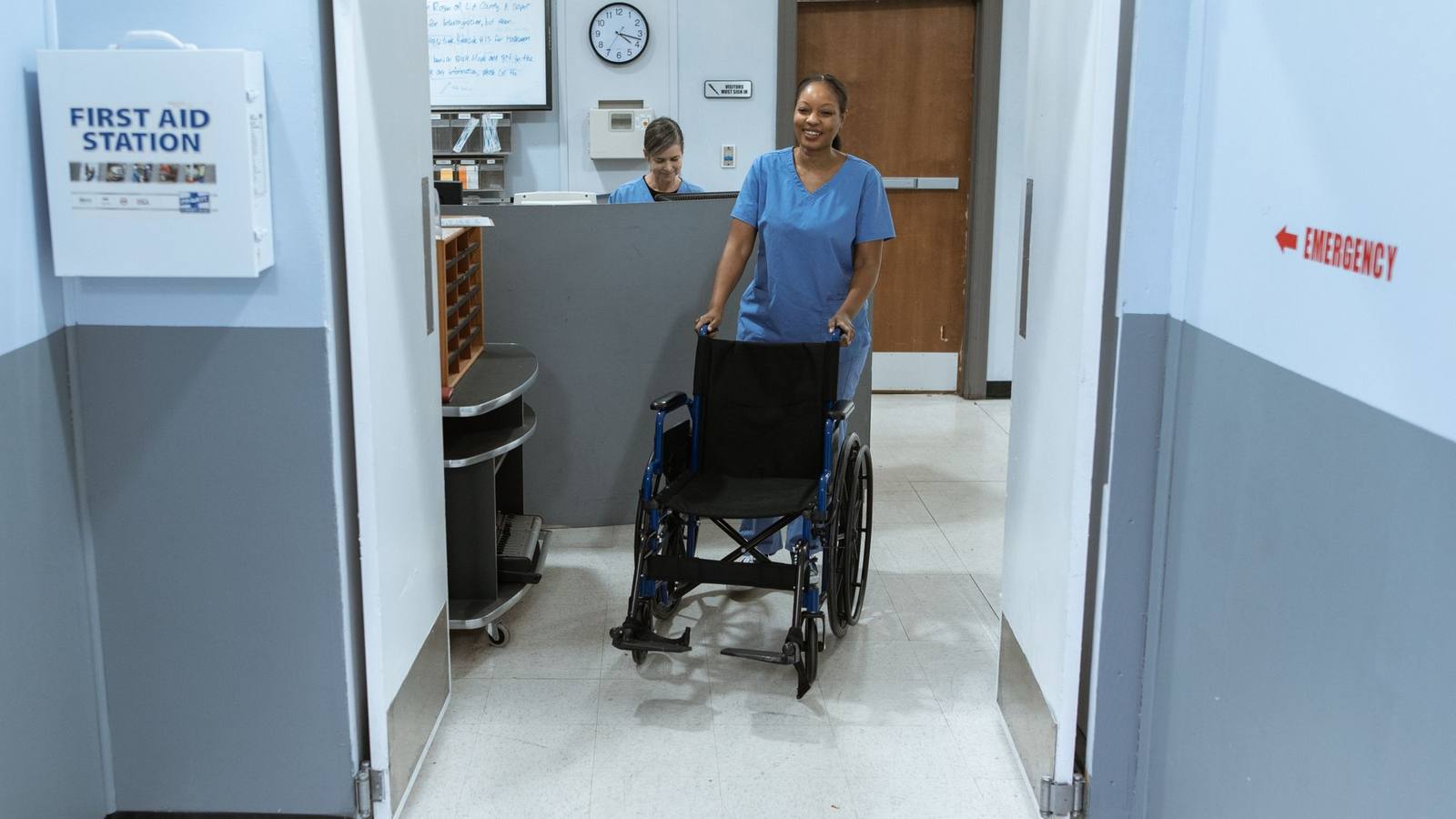 A nurse in scrubs pushing an empty wheelchair along a bright hospital corridor