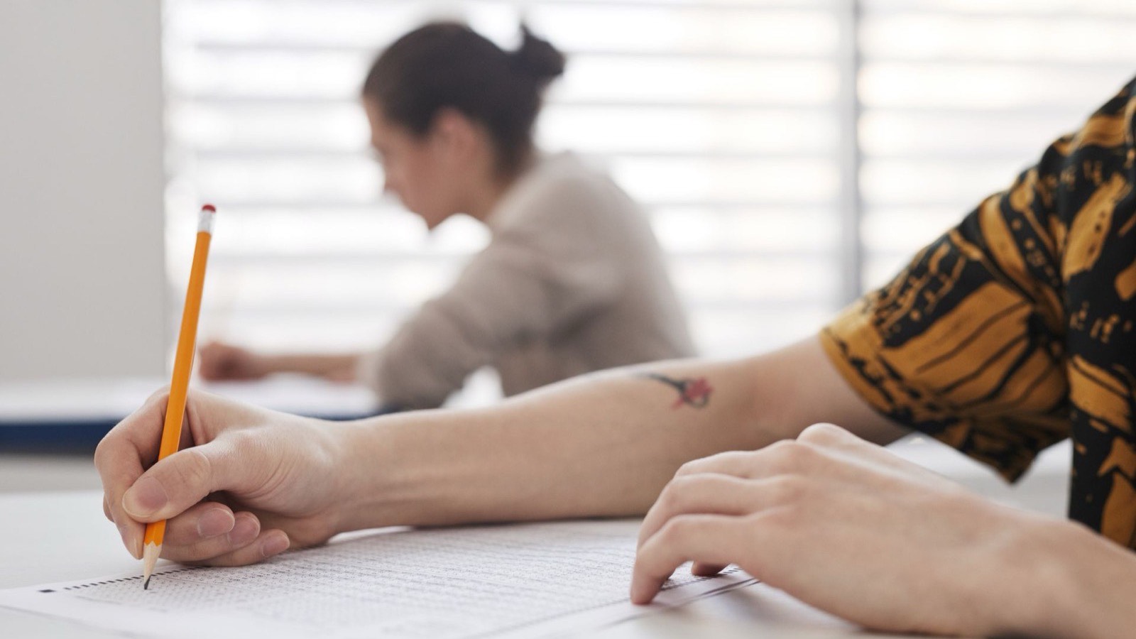 A hand holding an orange pencil fills in an answer sheet, with another test taker blurred in the background