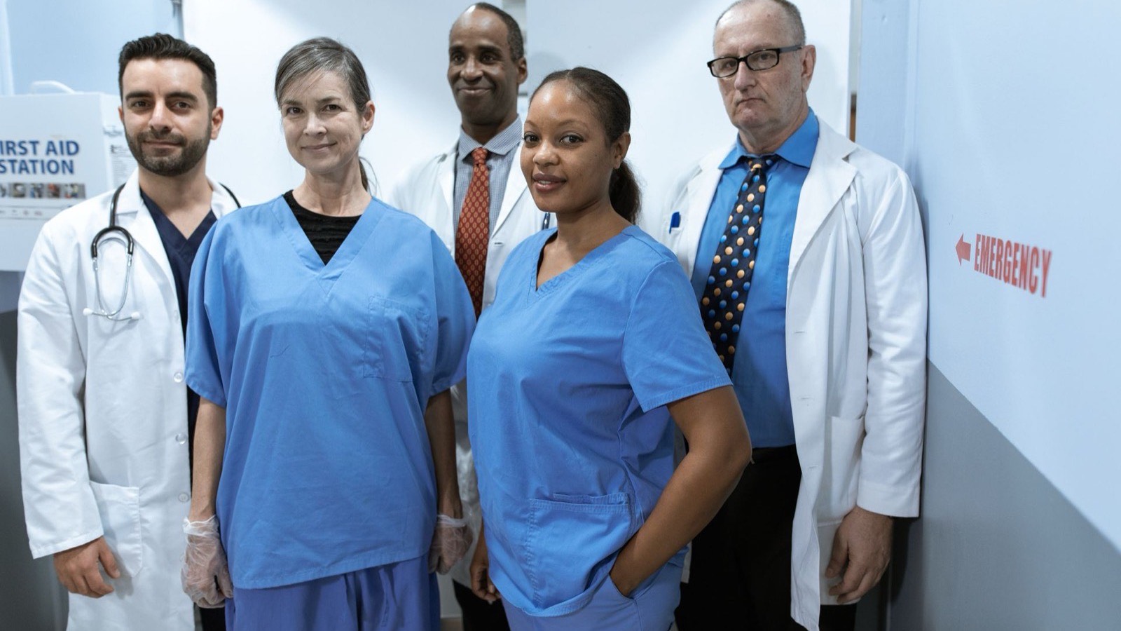 Two nurses in blue scrubs and three doctors in white coats standing in a hospital corridor next to an emergency sign